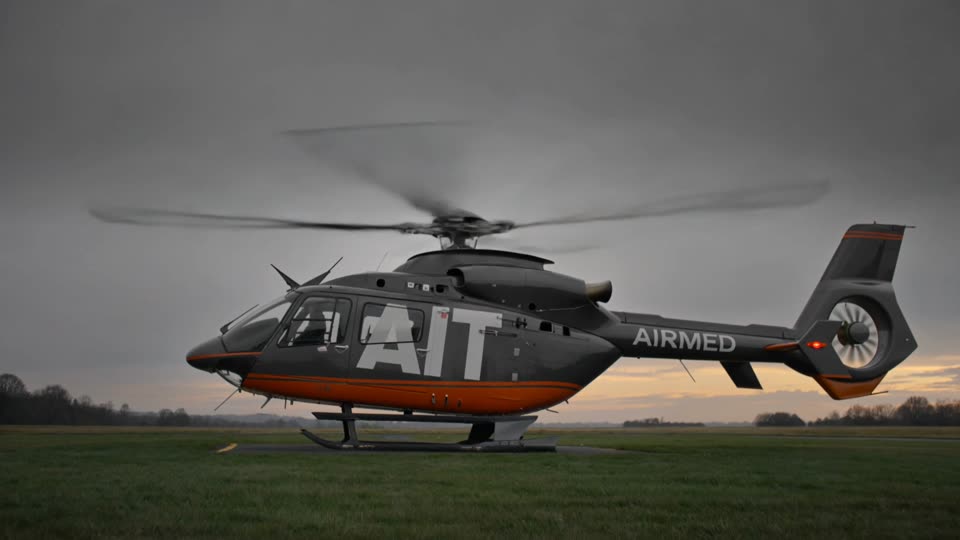 Medical helicopter with AIT on the fuselage and AIRMED on the tail boom, centered in frame at dusk, facing left, lifting off a grass helipad as rotor wash blasts grass and dust outward